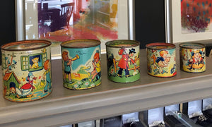 Five colourful tins of varying sizes placed on a shelf with framed pictures in the background.